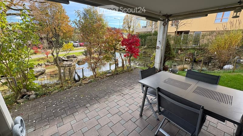 Balcony with dining table, black chairs, brick floor, view of a pond, trees, and greenery.
