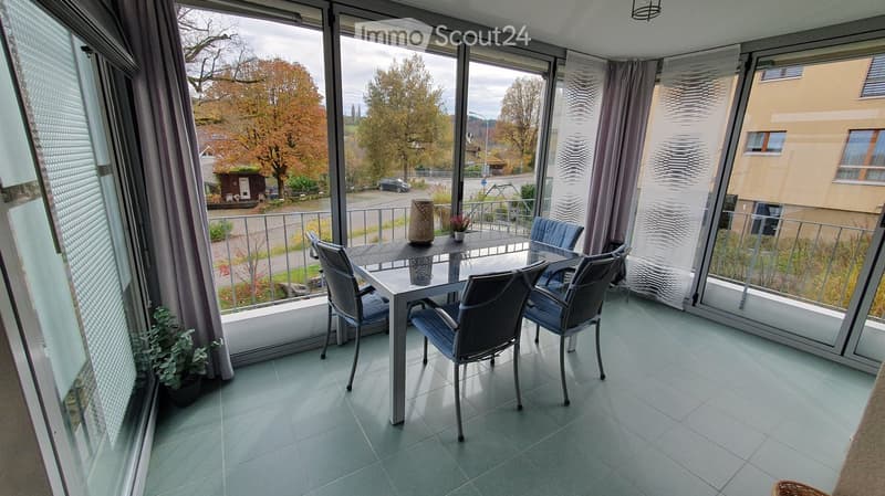 Table and chairs, balcony with glass railing, tiled floor, plant in a pot, outside view of street and trees