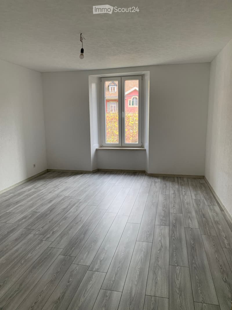 Empty room with light grey wooden flooring, white walls, and a large window with a view of a neighboring house.