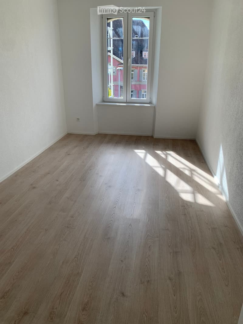 Empty room with light wooden flooring and white walls, featuring a large window with double doors and a radiator below it.