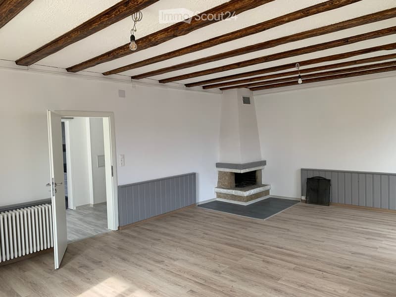 Empty room with wooden floor, exposed wooden beams, fireplace, and grey paneling on walls.
