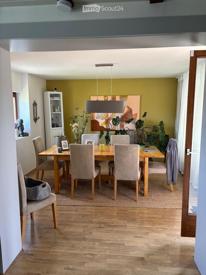Dining room with wooden floor, dining table with six chairs, pendant light, plants, picture frames, and a shelf with decorative items.