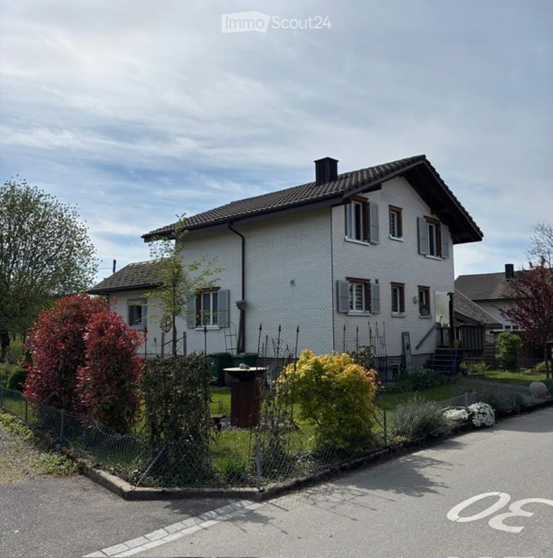 Two-story house with white exterior walls, multiple windows, and a dark roof. The house is surrounded by a small garden with various plants and trees.