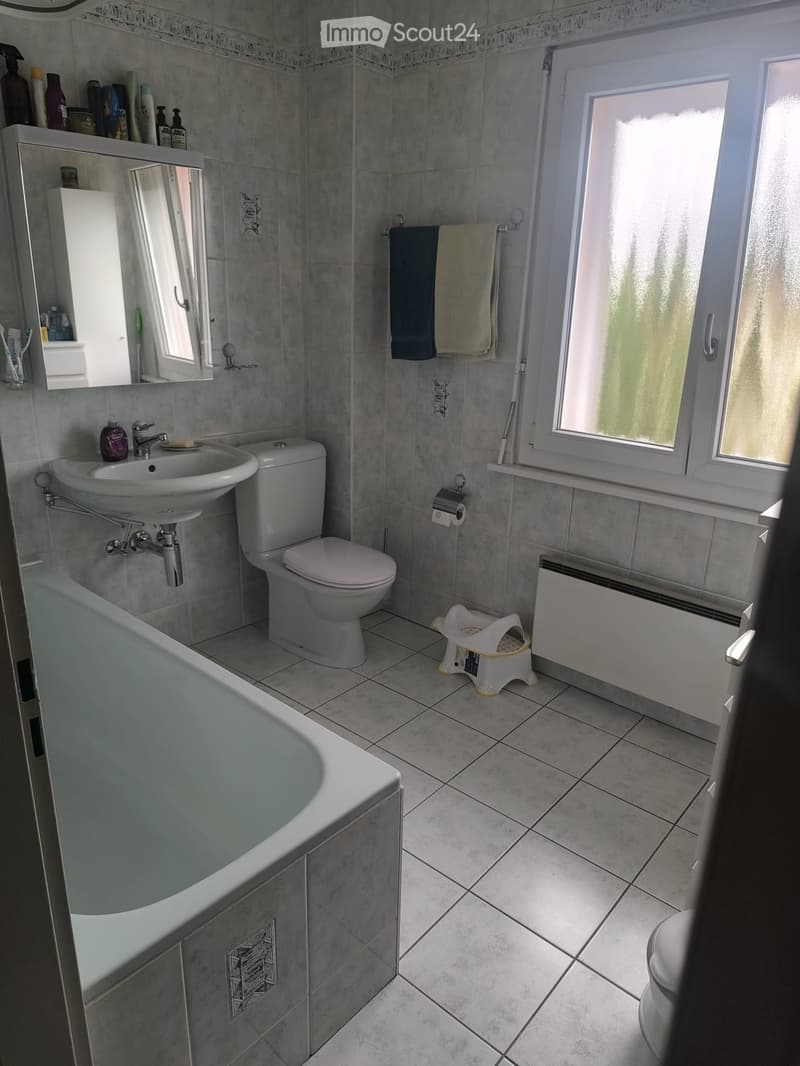 A modern bathroom with a bathtub, sink, toilet, mirror, and towel rack. The room features tiled walls and floor, a window with frosted glass, and a small white stool near the radiator.