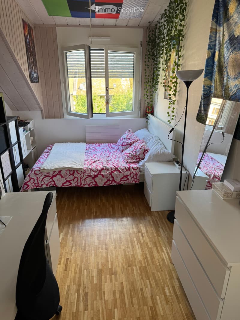 Cozy bedroom with wooden flooring, a single bed with pink and white bedding, a nightstand, a mirror, a chair, and a floor lamp. The room features a large window with blinds and a view of greenery outside.