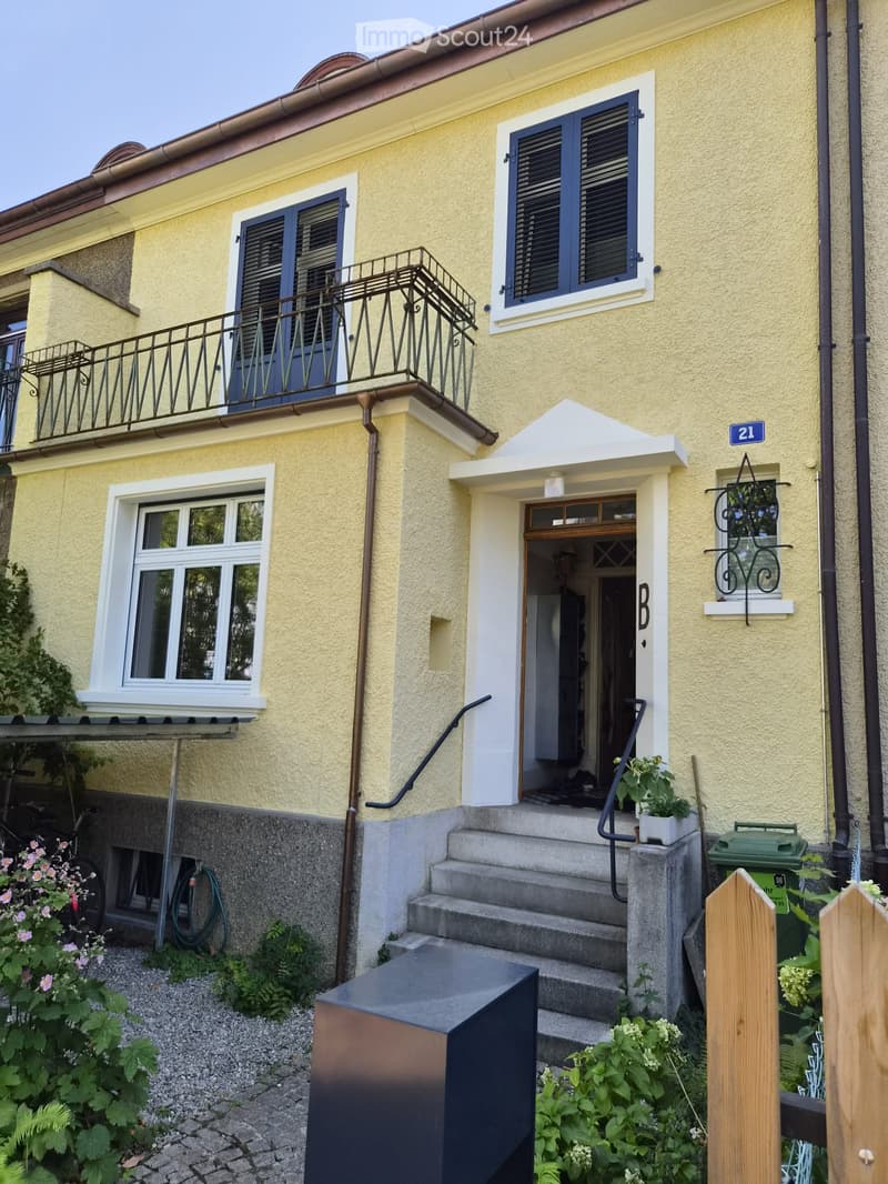 Two-story yellow building with white trim, blue shutters, and a small balcony on the upper floor. The entrance features a small porch with steps and a potted plant. The house number "21" is displayed on the wall.