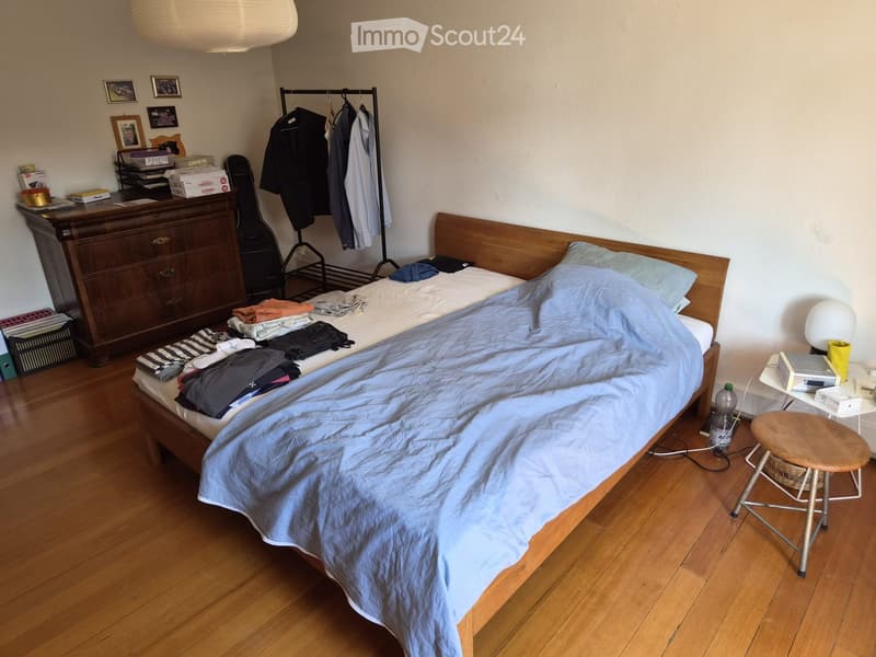 A small bedroom with a wooden bed, a wooden stool, a guitar, a lamp, a table, and a cup on a small table. The bed is unmade with clothes and a blanket on it.