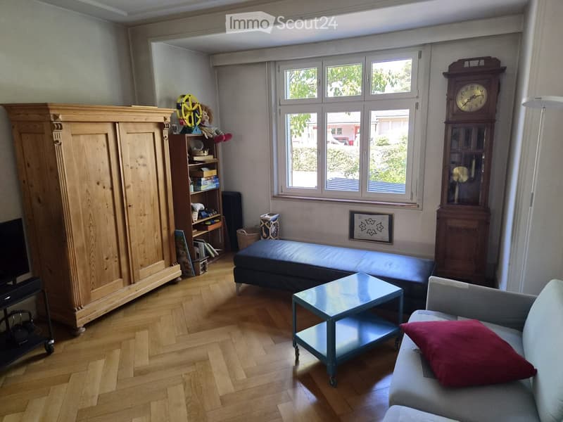 Living room with wooden floor, large window, grey sofa with red pillow, coffee table, armchair, cabinet, bookshelf, grandfather clock, ottoman, and screen.