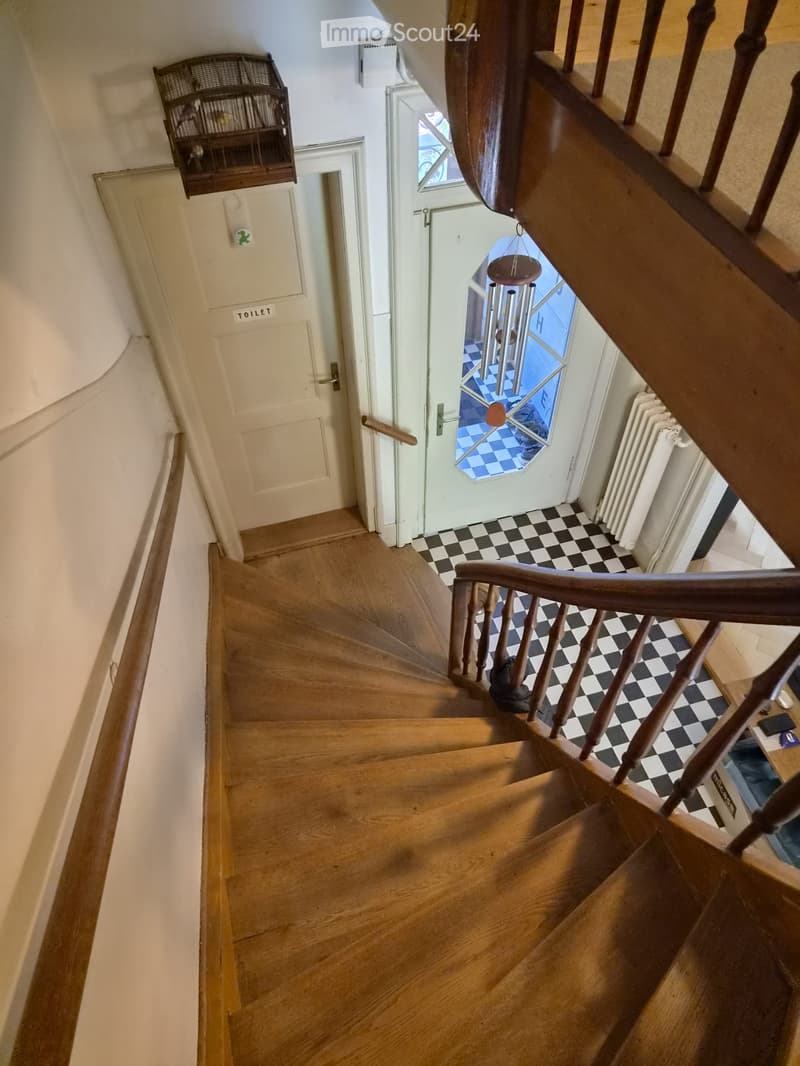 Spiral staircase with wooden steps and handrail, checkered floor, white walls, a door labeled 'TOILET', and a hanging birdcage visible on the wall.