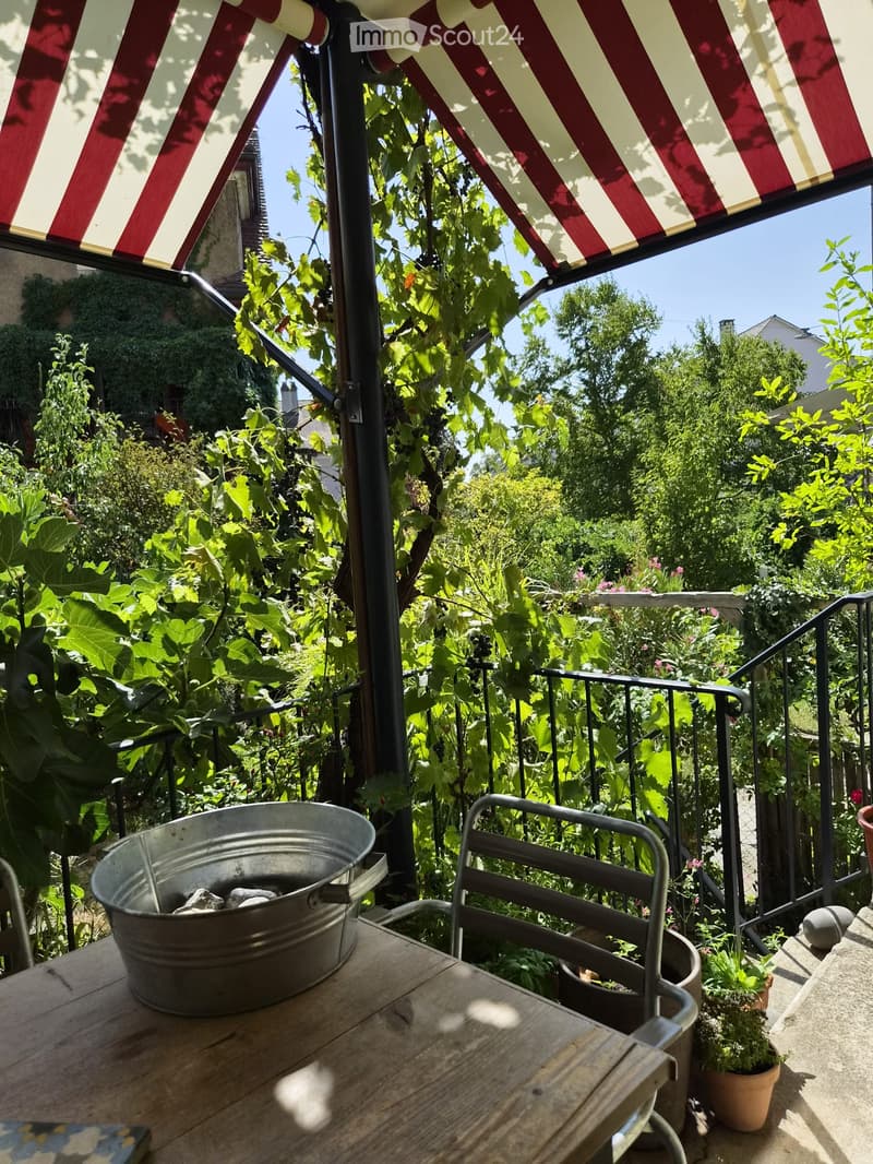 Balcony with wooden table, metal chair, metal bucket, and potted plants, surrounded by lush greenery and a red-and-white striped awning.