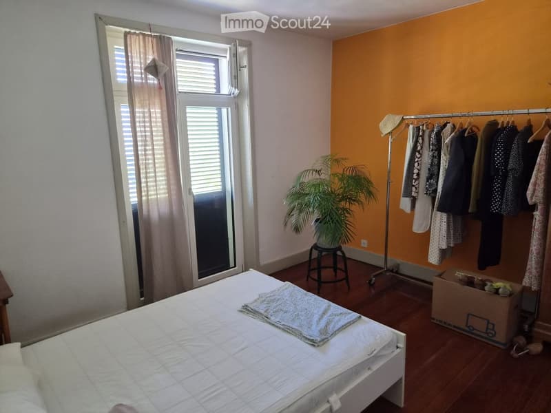 Bedroom with a white bed, a small stool with a plant, a clothing rack with hanging clothes, and a large window with blinds and a curtain.