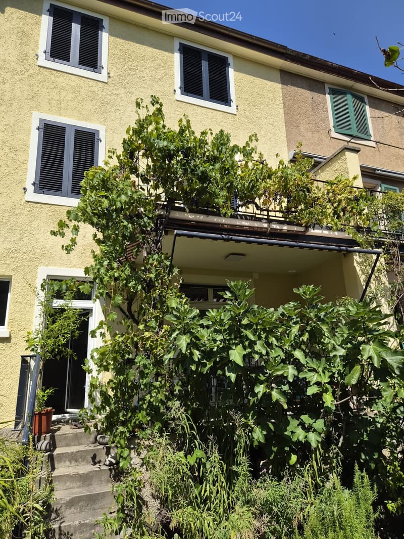 A lush garden area in front of a two-story house with yellow walls and white-framed windows, featuring climbing vines and various plants.