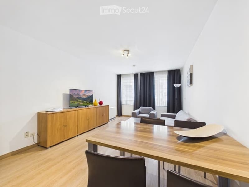 Modern living room with a sofa, coffee table, TV, dining table, chairs, and a wooden cabinet. The room features large windows with dark curtains and a sleek design.