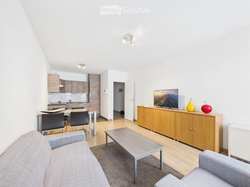 Modern open-plan living and dining area with a grey sofa, coffee table, dining table with chairs, kitchen cabinets, and a television on a sideboard with decorative fruits.