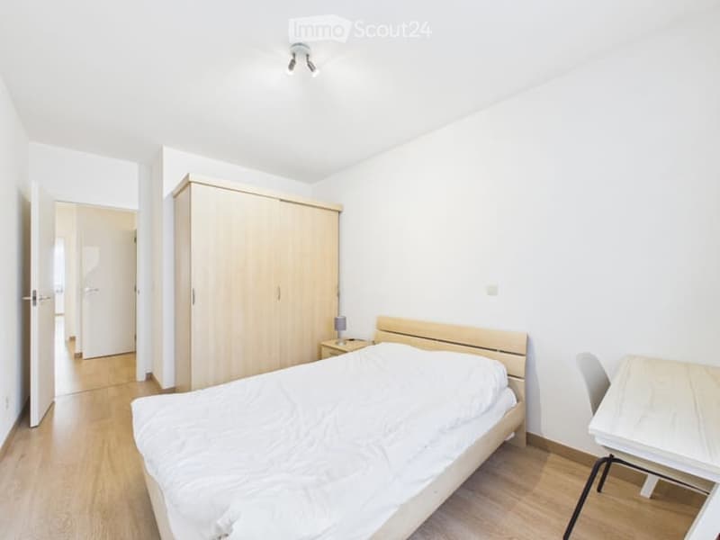 Bright bedroom with white walls, wooden floor, bed with white bedding, wardrobe, nightstand, small table, and chair.