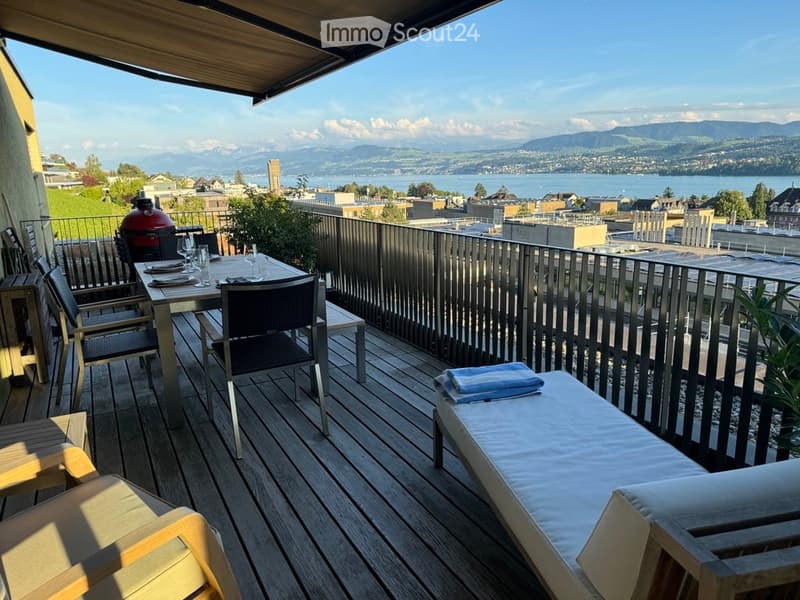 Balcony with wooden decking, outdoor dining set, sunbed, and grill, overlooking a scenic lake and mountain view.