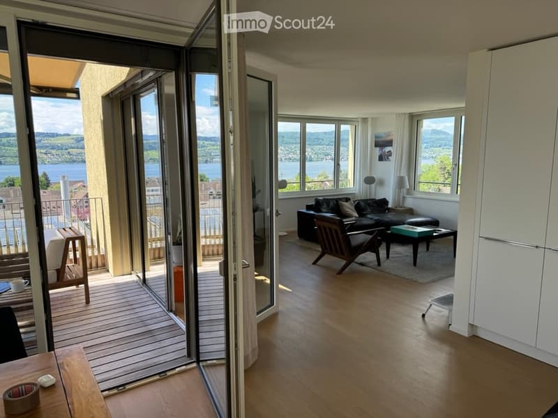 Bright living room with large windows and a balcony overlooking a scenic lake view. Features include a black leather sofa, a wooden coffee table, a wooden chair, and a small table with a cup on it.