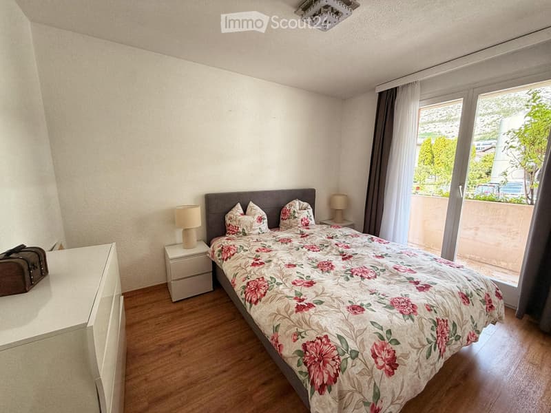 A well-lit bedroom with a wooden floor, a large bed with floral bedding, nightstands with lamps, and a view of a balcony through large windows.
