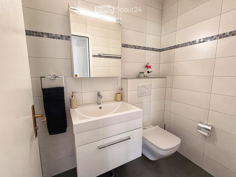 Modern bathroom with white tiles, a white sink with a modern faucet, a large mirror with built-in lighting, a toilet, a towel, and a soap dispenser.