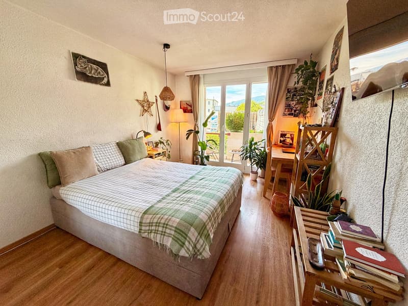 Cozy bedroom with a bed, nightstand, desk, and balcony access. Features wooden flooring, modern decor, and a view of the outdoor area.