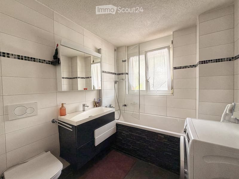 Modern bathroom with white tiles, a large mirror, a sink with a dark cabinet, a bathtub, and a toilet. The space features a window with frosted glass and a towel hanging on the side.