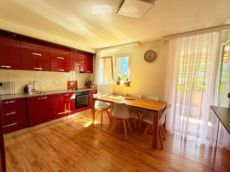 Modern kitchen with red cabinets, wooden floor, and a dining area with a table and chairs. Features include a sink, oven, dishwasher, and a window with sheer curtains.