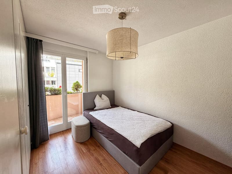 Cozy bedroom with a single bed, white pillows, a round stool, and a pendant light fixture. The room features wooden flooring and a large window with a curtain leading to a small balcony.