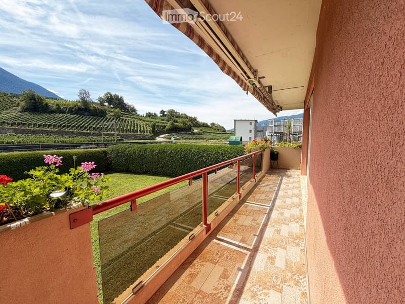 Balcony with tiled floor, glass railing, and flower pots, overlooking a landscaped garden and vineyard area with mountains in the background.