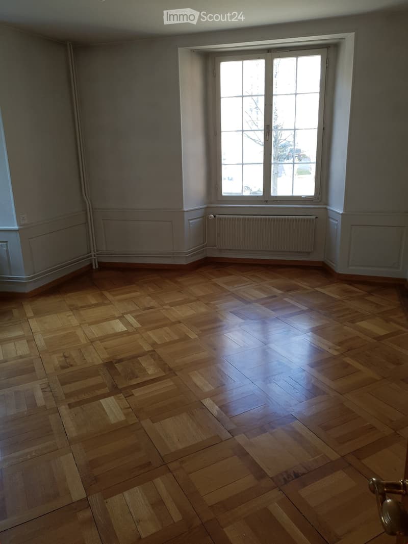 Empty room with wooden parquet flooring, white walls, and large windows with white frames. Radiators are visible along the walls.