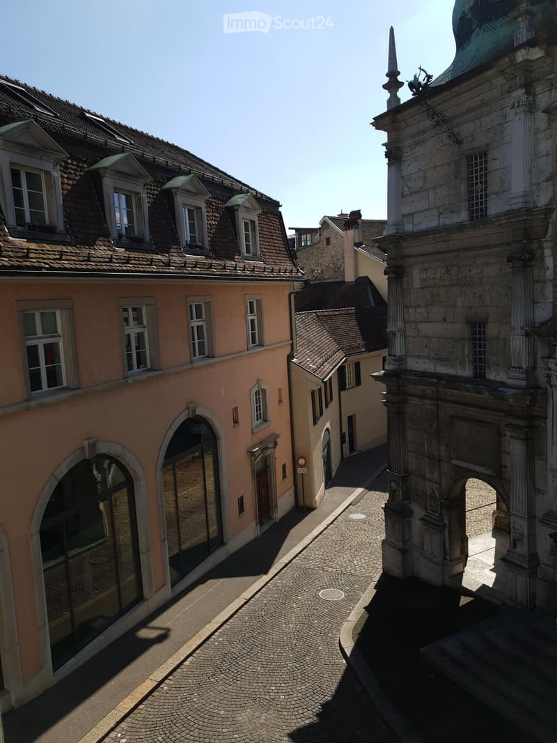A narrow, cobblestone alleyway flanked by historic buildings with varied architectural styles, including a peach-colored building with large windows and a more ornate stone structure with a spire.