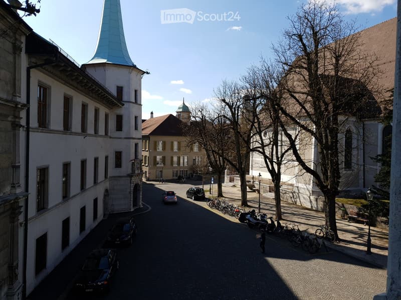A narrow cobblestone street lined with historic buildings, including a tall tower with a blue spire and a church with a green dome. Several parked cars and bicycles are visible, along with a few pedestrians.