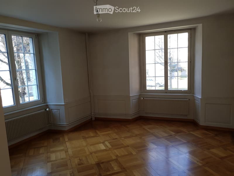 Empty room with wooden flooring, large windows, radiators, and a hanging light fixture.