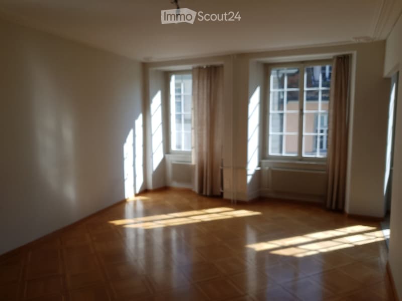 Empty room with wooden flooring, large windows with curtains, and radiators beneath the windows.