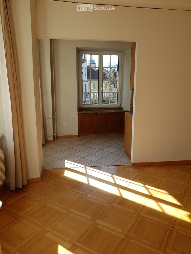 Empty room with wooden flooring and tiled section, featuring a window with a view of a building and a small cabinet area.