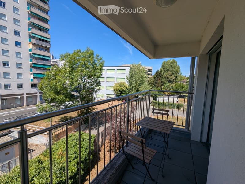 Balcony with a small wooden table and two chairs, overlooking a residential area with trees and buildings.