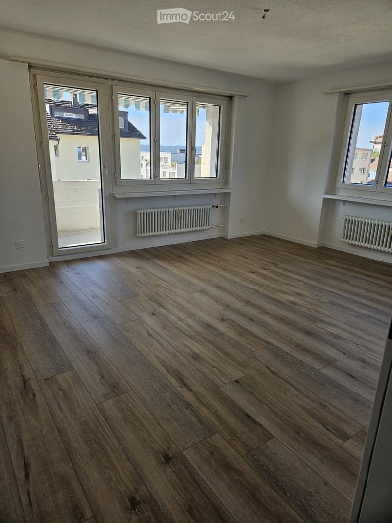 Empty room with wooden flooring, large windows, radiators, and a sliding glass door leading to a balcony.