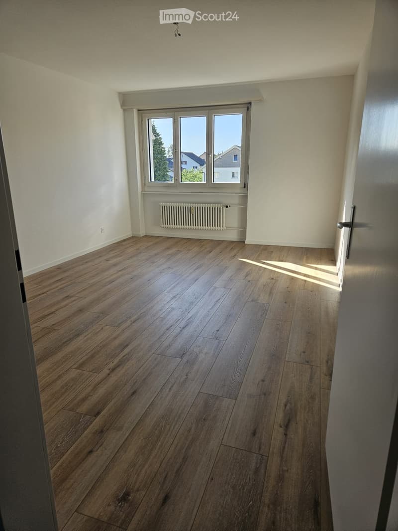 Empty room with wooden flooring, white walls, large window with blinds, radiator, and a door.