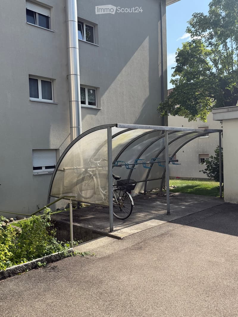 Modern building exterior with a covered bicycle parking area, featuring a sleek glass canopy and metal frame, with a bicycle parked inside.
