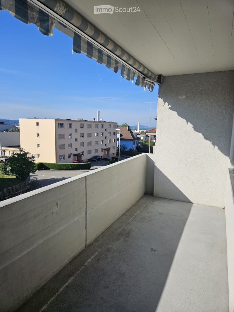 Balcony with concrete floor, low wall, striped awning, overlooking a parking lot and residential buildings.