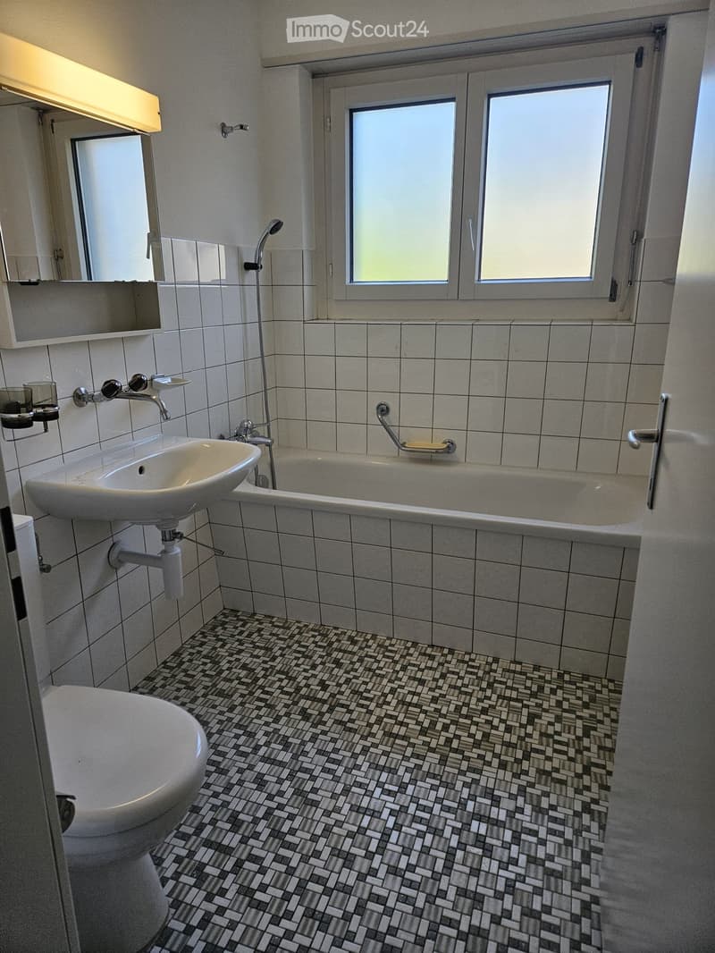 Modern bathroom with white tiles, a bathtub, a toilet, a sink, and a showerhead. The floor features a patterned tile design, and there is a large window providing natural light.