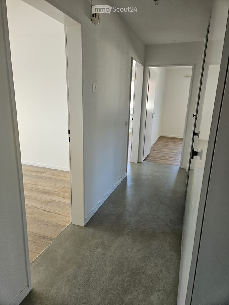 Empty hallway with light grey walls, polished concrete floor, and wooden flooring visible in adjacent rooms. Features a smoke detector and electrical outlet on the wall.