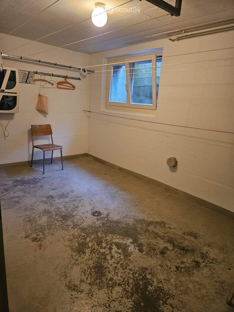 Empty room with concrete floor, single window, clothes rack, and a single chair. Bare walls and minimal furnishings.