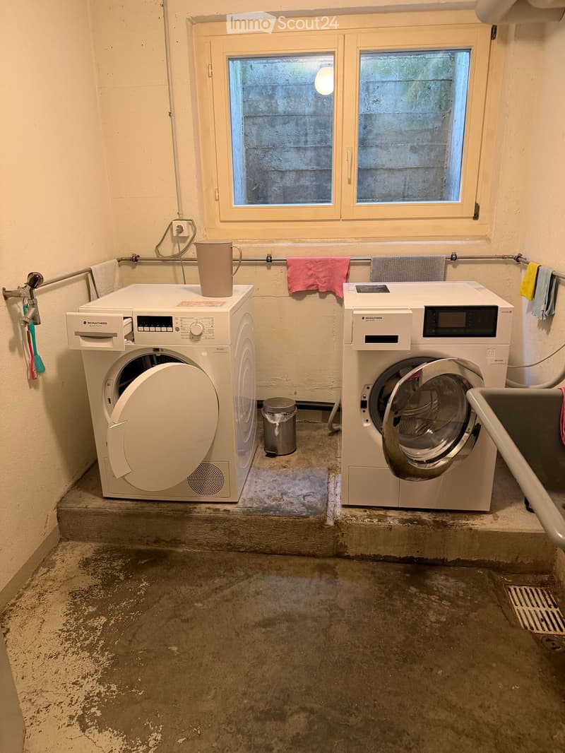 Laundry area with a washing machine, dryer, and a cup on top of the washing machine. Towels are hanging on a rack, and a small trash can is placed between the machines.