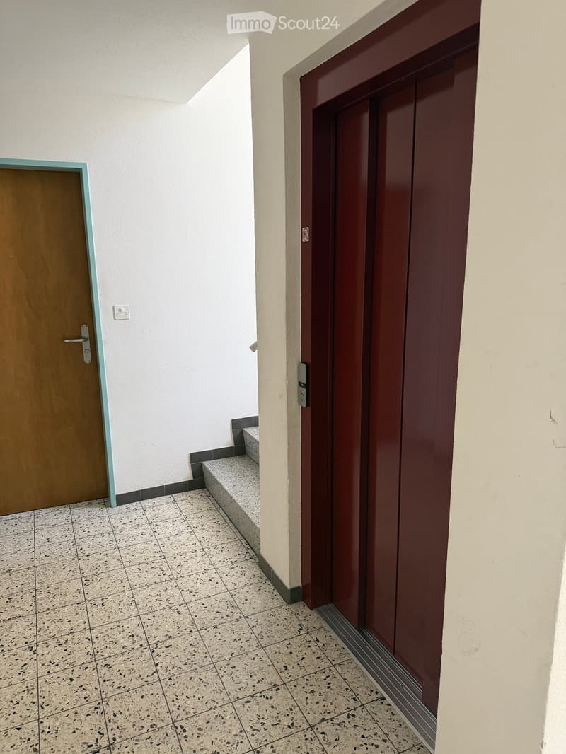 Hallway with tiled floor, two doors (one wooden and one metallic), and a small bench in the corner.