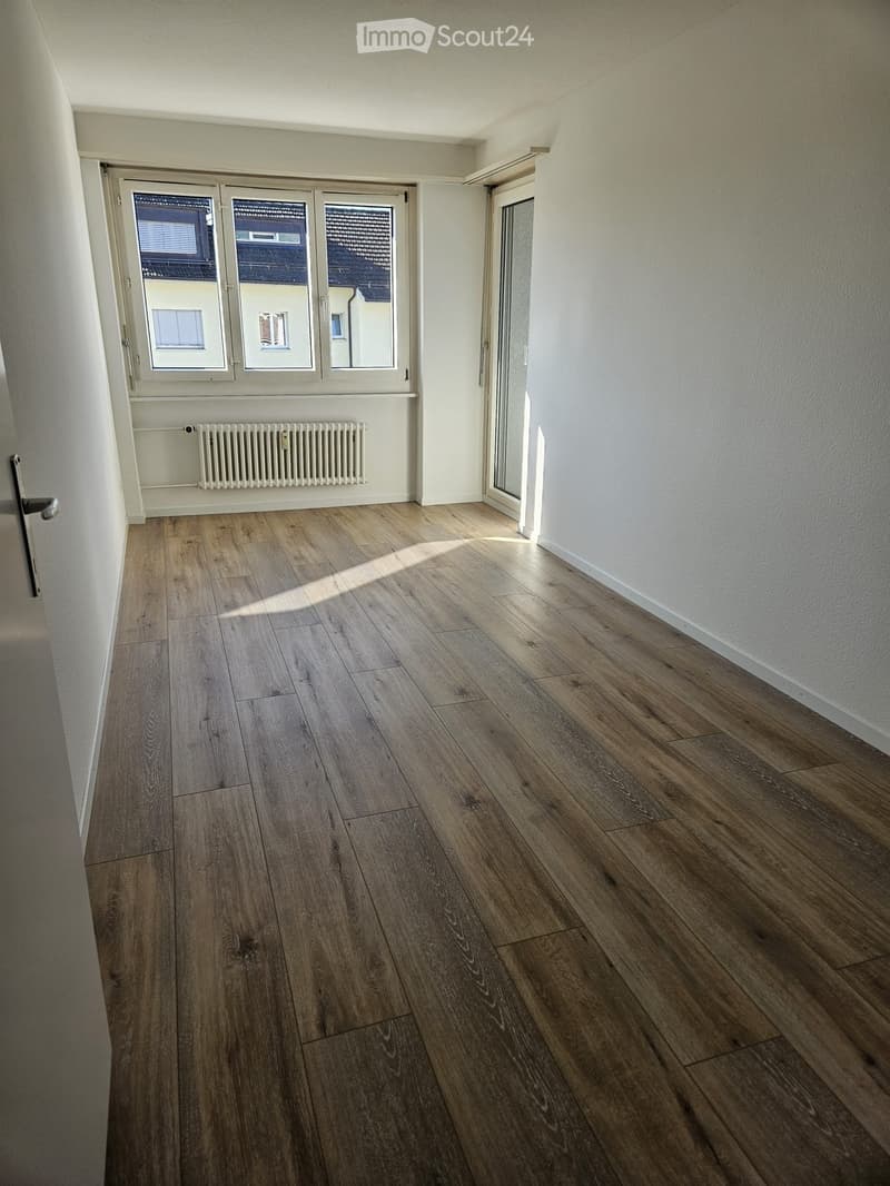 Empty room with wooden flooring, white walls, large windows, and a radiator. No furniture or equipment present.