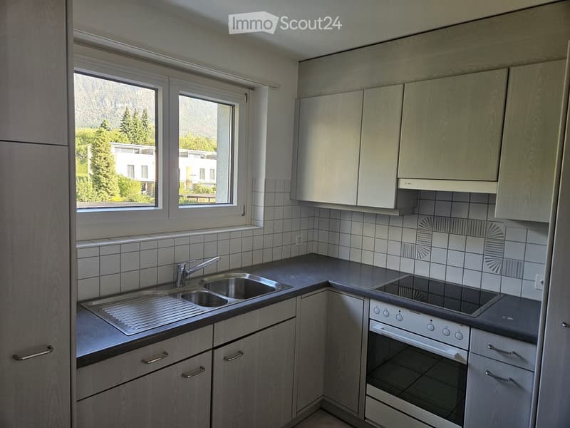 Modern kitchen with light wood cabinets, stainless steel sink, faucet, oven, and stove. The kitchen features tiled walls and large windows offering a view of greenery and buildings.