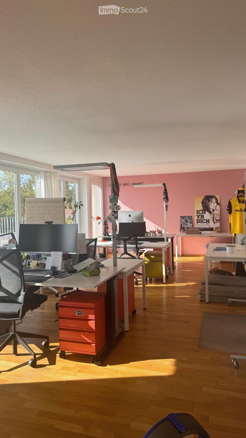 Brightly lit office space with wooden flooring, multiple desks, chairs, and various office equipment including screens, laptops, and a cup on a table. There is a poster on the wall and a small plant near the window.