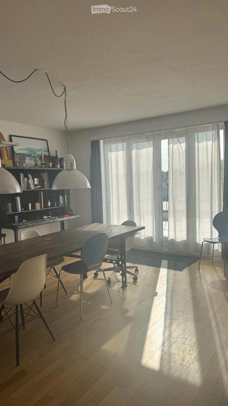Bright dining area with wooden floor, large dining table, modern chairs, pendant lamps, and a shelf with books and decorative items.