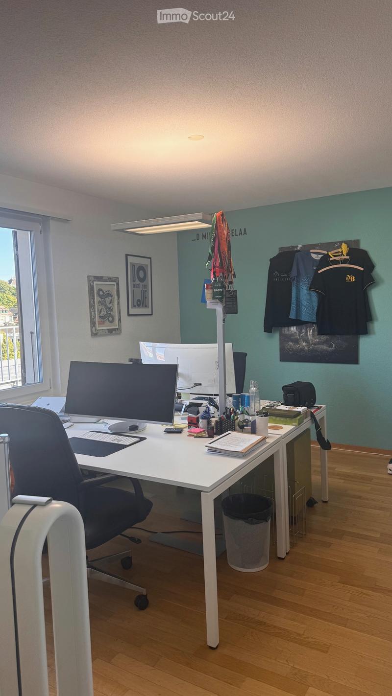 A modern, well-lit office space with a white desk, ergonomic chair, computer setup, and various office supplies. The room features light wood flooring, a large window with a view, and decorative elements including framed artwork and t-shirts hanging on th