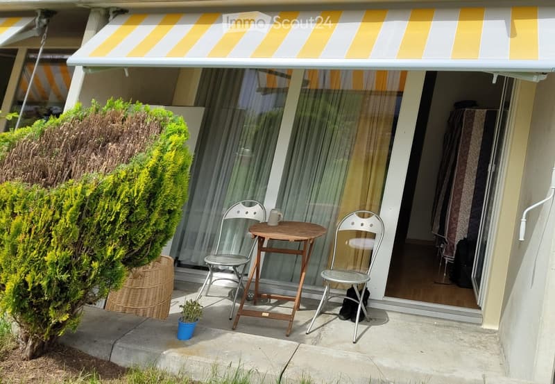 Balcony with a small round wooden table, two white metal chairs, a cup, and a small potted plant. A large sliding glass door leads to the interior, and a yellow and white striped awning provides shade.